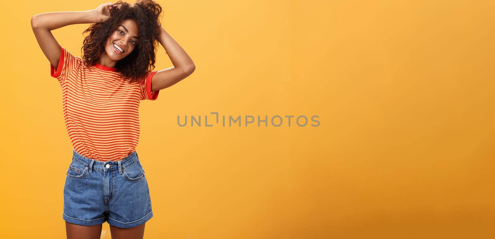 Time start living life fullest. Joyful optimistic woman having fun during vacation tilting head touching curly hair and enjoying summer sunshine in trendy striped t-shirt and shorts over orange wall by Benzoix