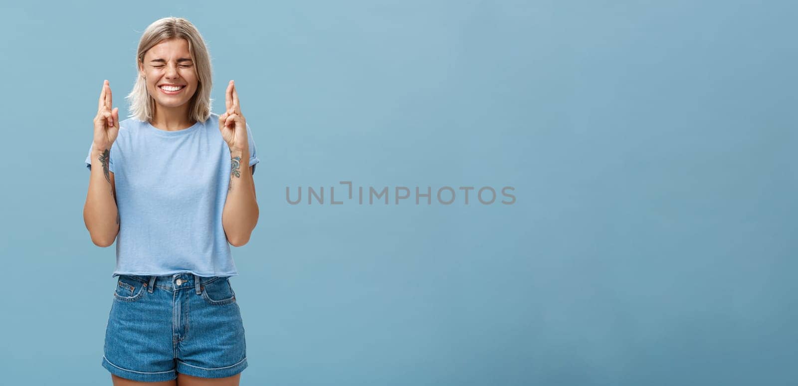 Optimistic faithful good-looking young woman with blond hair and tattoos smiling joyfully crossing fingers for good luck waiting for dream come true making wish while standing over blue background. Copy space