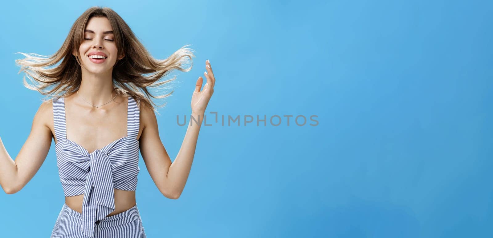 Woman feeling hair filled with strengths after washing with new shampoo or cosmetology product playing with hairstyle closing eyes with delighted happy smile standing tender and sensual over blue wall. Treatments, cosmetics, hairdressing concept