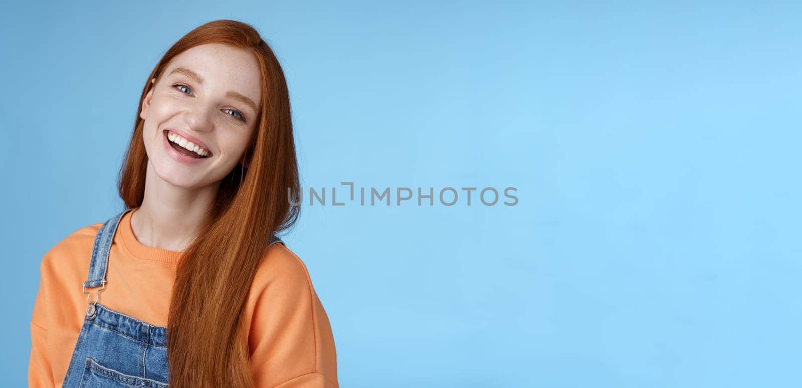 Positive outgoing lively redhead girl laughing joyfully having fun talking friendly friends tilting head chuckling joking funny life moments standing positive lucky blue background orange t-shirt by Benzoix