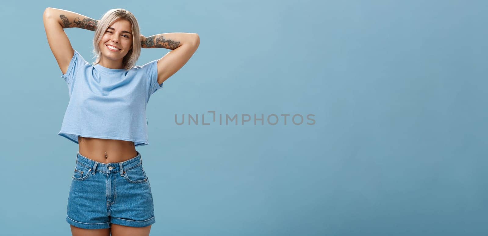 Thanks god it weekends. Relaxed lazy and attractive young stylish woman with fari hair tattoos, pierced belly stretching with delight holding hands behind head in carefree pose, smiling over blue wall.