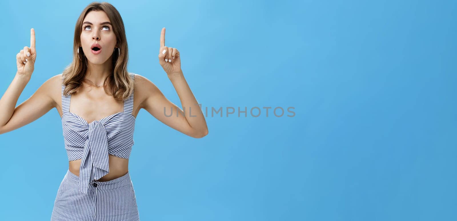 Waist-up shot of curious good-looking stylish modern woman dropping jaw saying wow while lifting head up looking and pointing upwards with amazed and surprised expression posing against blue background. Copy space