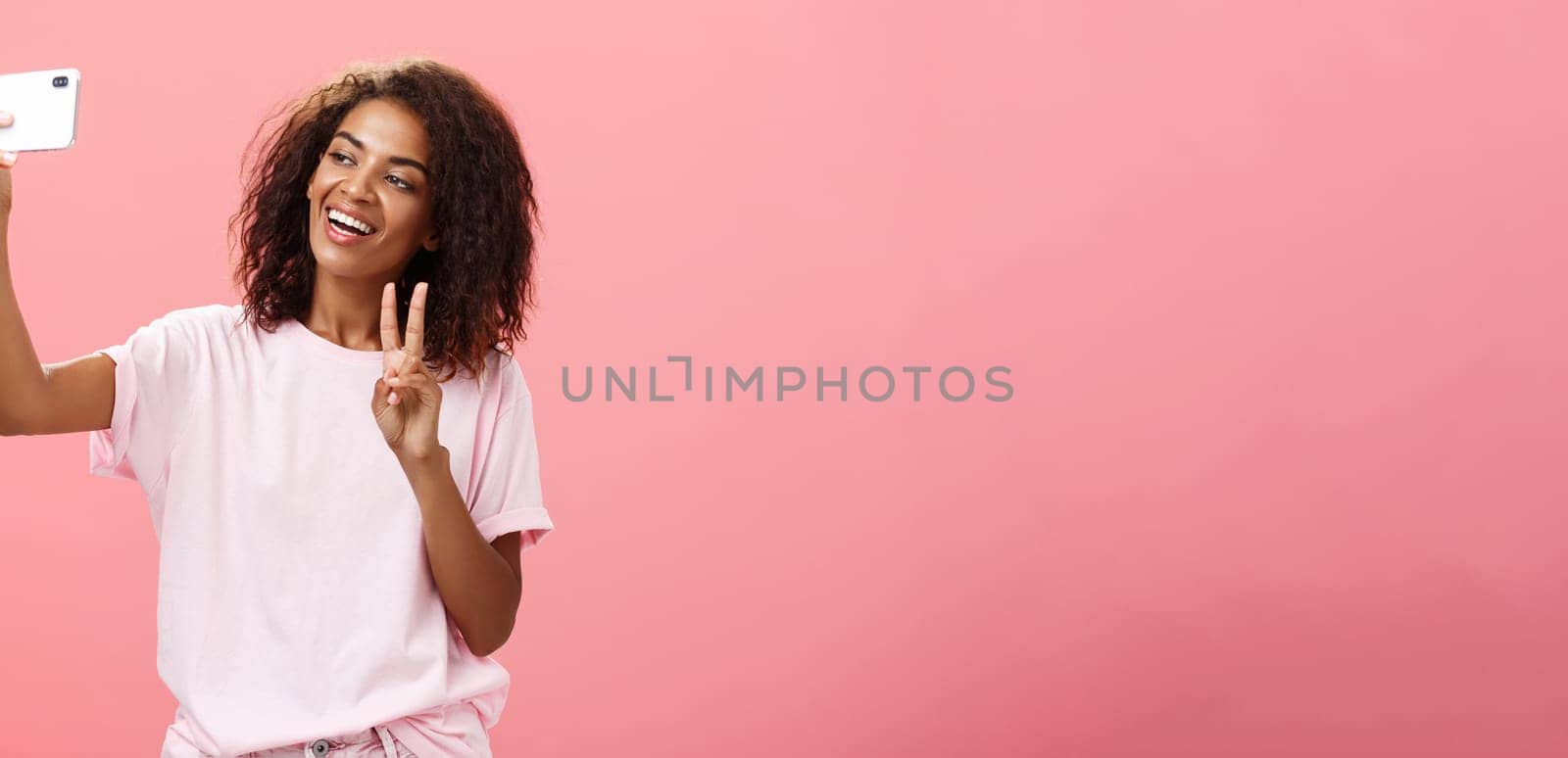 Stylish sociable good-looking dark-skinned female student with curly hairstyle pulling hand with smartphone near face taking selfie showing peace sign to device screen while smiling carefree by Benzoix