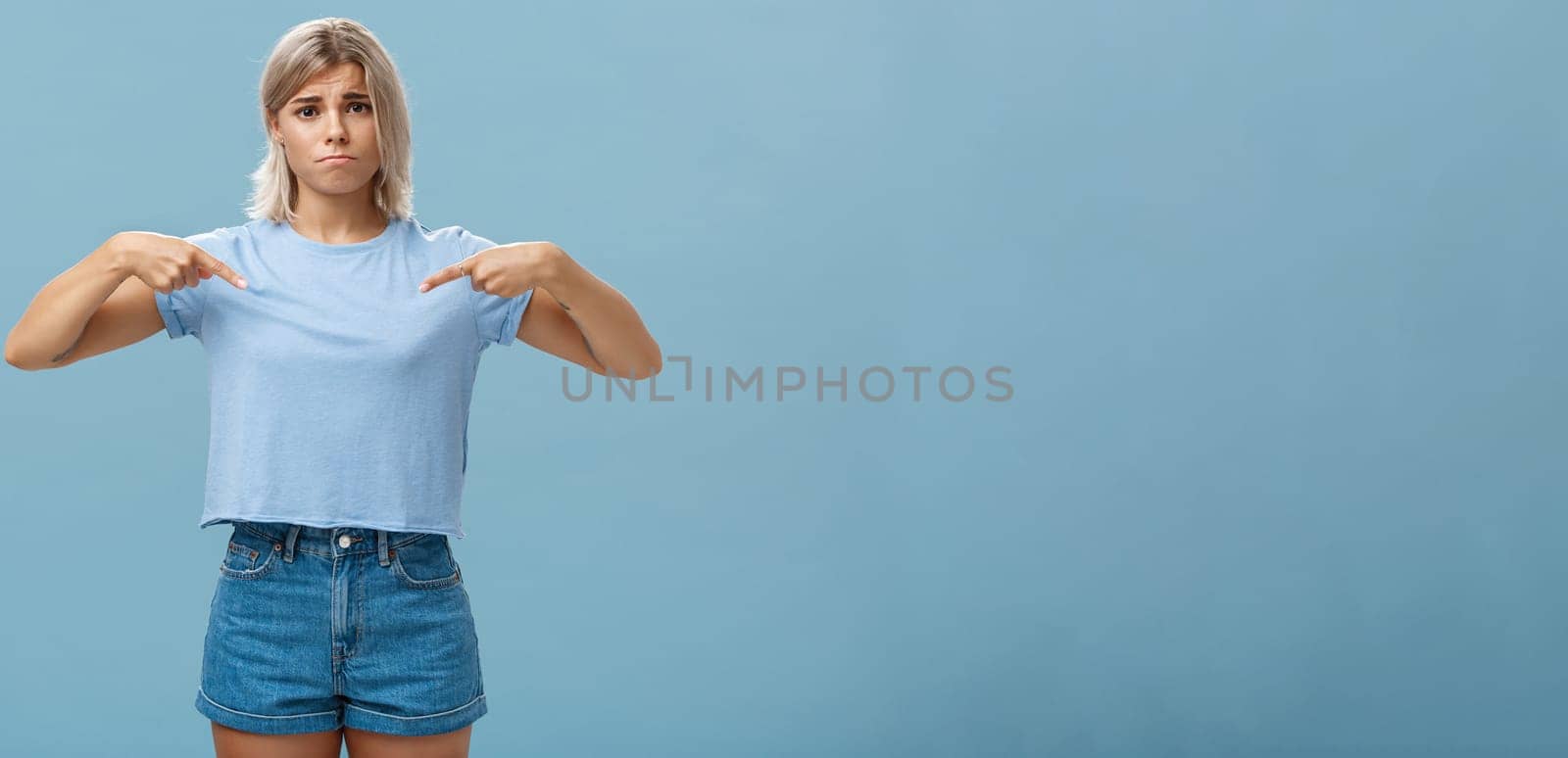 Sad cute blonde young female complaining being displeased with own body, pointing at breast frowning making gloomy smile pulling lips down from disappointment standing over blue background.