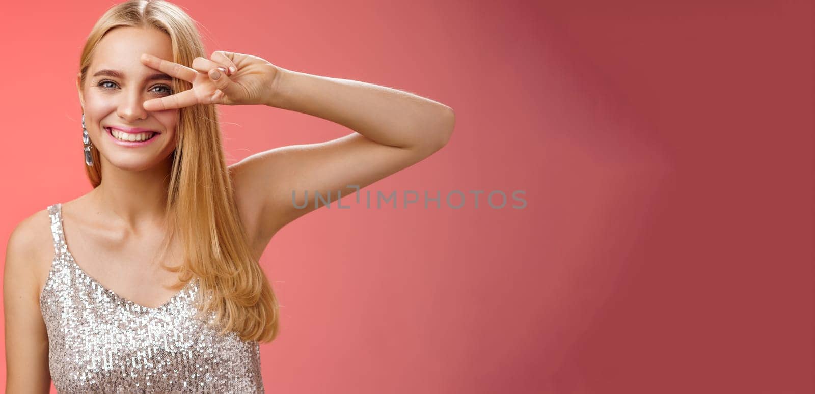 Lifestyle. Joyful carefree attractive confident blond woman in stylish silver dress show disco sign victory peace gesture near eye smiling having fun enjoying awesome party grinning dancing red background.