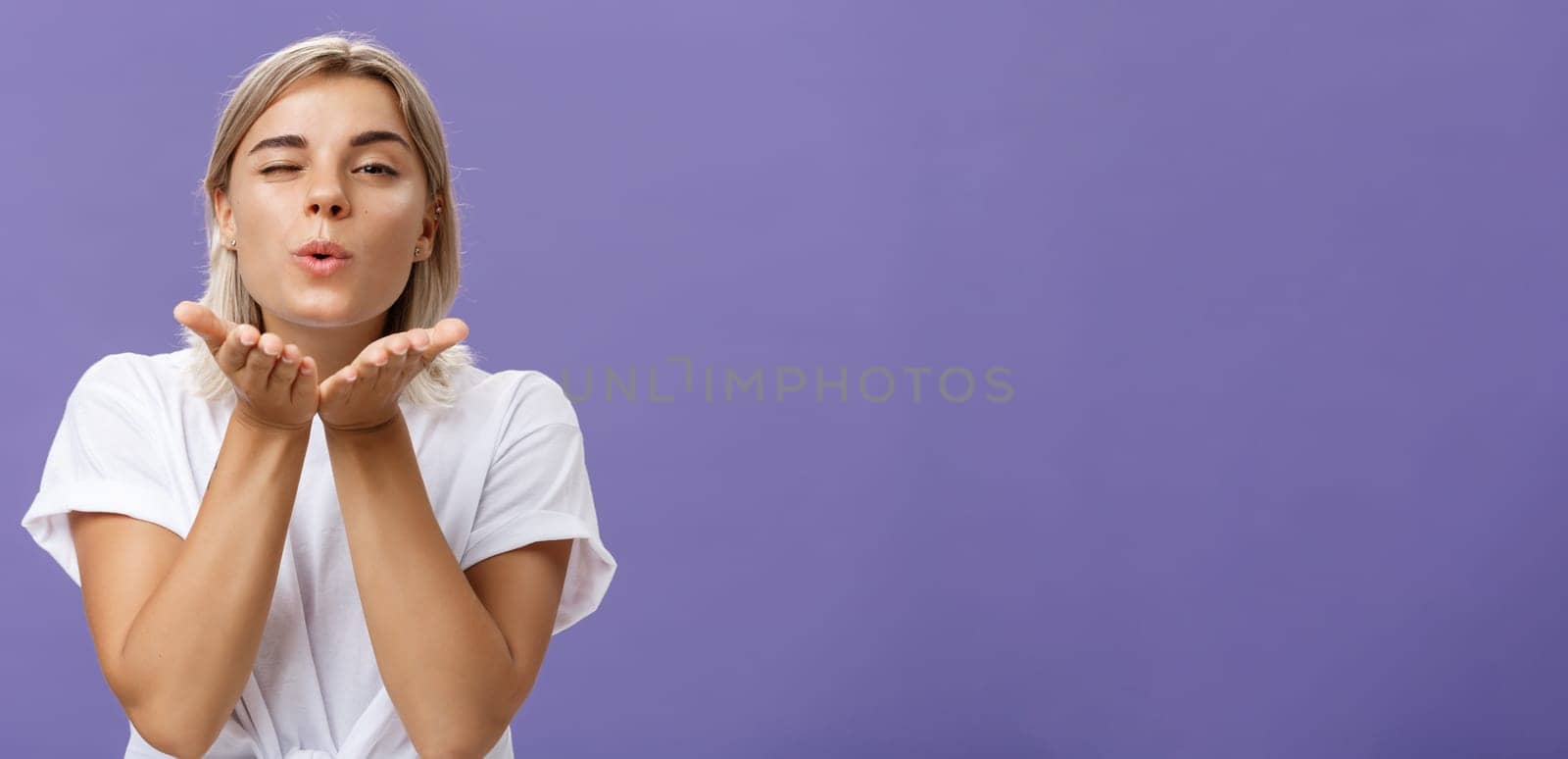 Blowing sweet kiss to all fans. Flirty tender and cute stylish european female with blond hair and tanned skin holding palms near mouth folding lips while sending mwah at camera over purple wall.