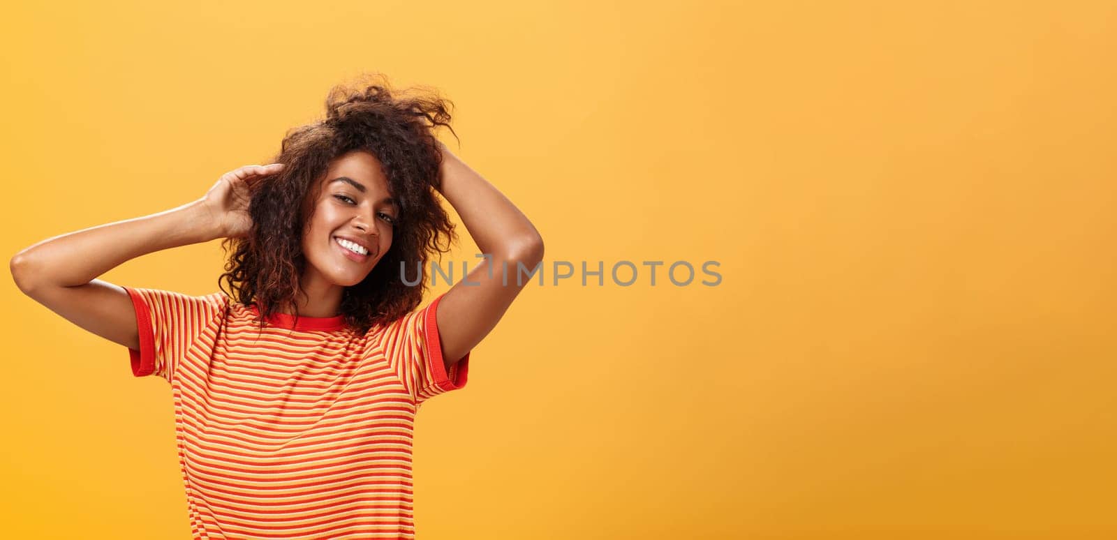 Waist-up shot of charming flirty feminine dark-skinned female in playful mood dancing playing with curly hair and smiling with delight and joy posing over orange background happy and carefree by Benzoix