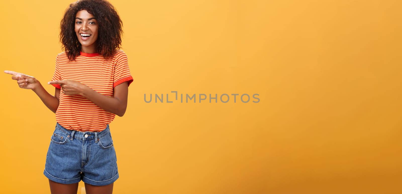 Ask him. Portrait of friendly and joyful good-looking stylish female shop assistant with curly hair and dark skin pointing left with both hands, smiling assured and entertained over orange background by Benzoix
