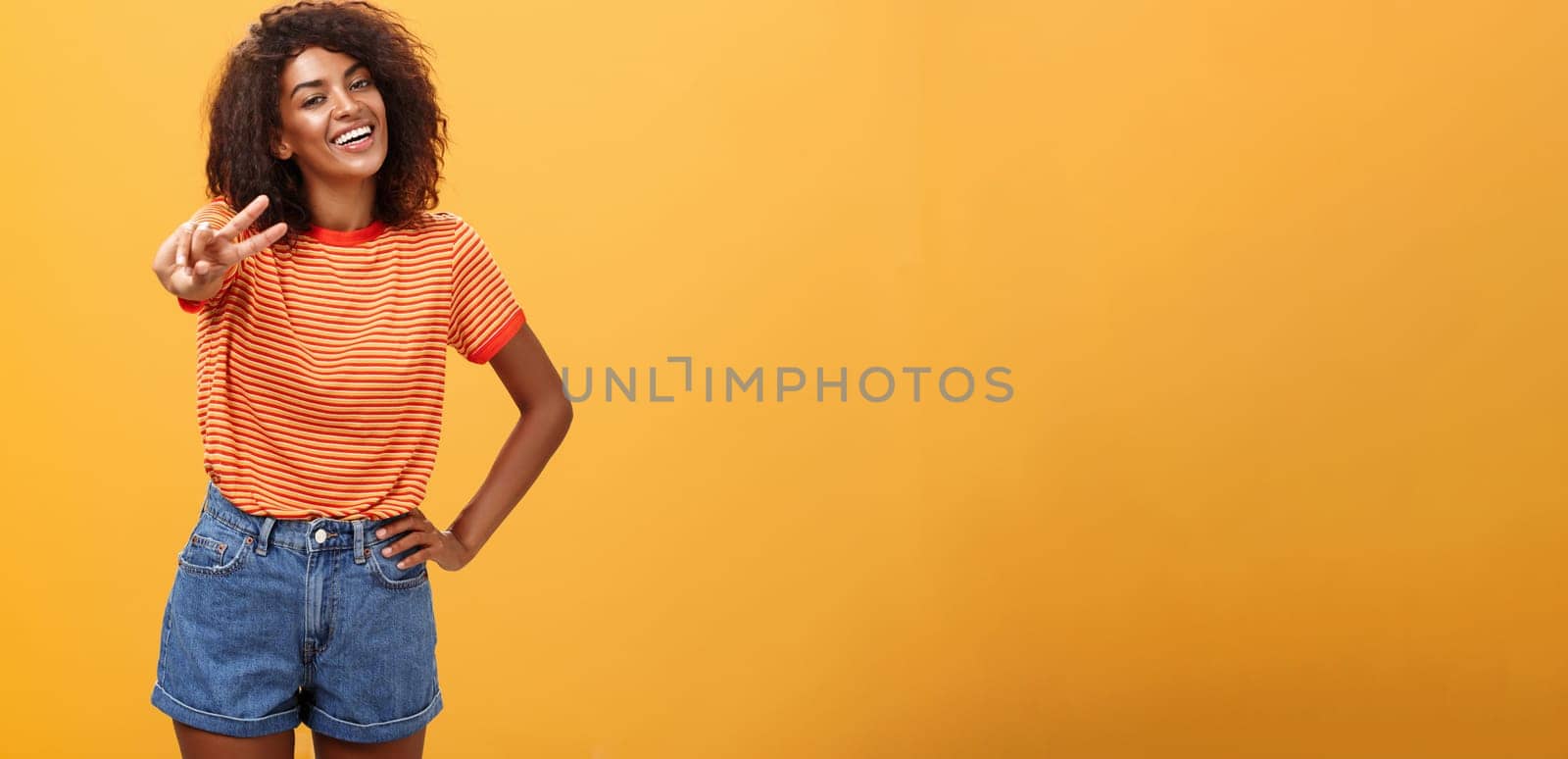 Friendly carefree and happy good-looking tall african american female model with curly hairstyle holding hand on hip tilting head and smiling joyfully showing peace gesture over orange wall. Copy space