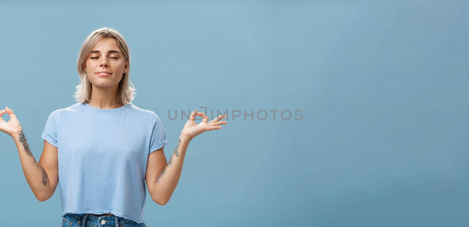 Listening to inner self during meditation. Calm happy and relaxed attractive tanned blonde female with tattoos on arms standing in lotus pose over blue wall with zen gesture closed eyes doing yoga by Benzoix