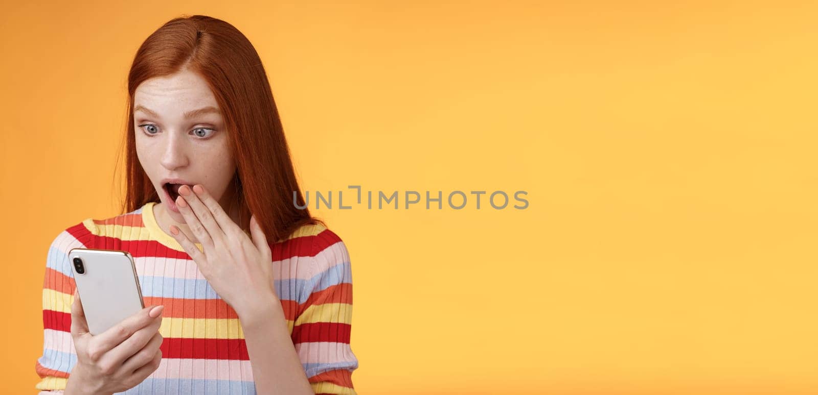 Amazed speechless young teenage redhead girl student gasping drop jaw say omg wow cover opened mouth palm look shocked surprised smartphone display reading fresh gossips orange background by Benzoix
