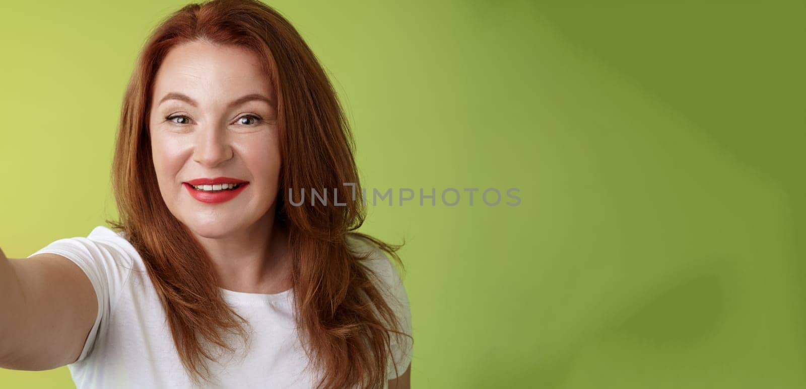 Close-up joyful enthusiastic redhead alluring middle-aged woman extand arm towards camera taking selfie smartphone smiling broadly posing photograph taking picture device green background.