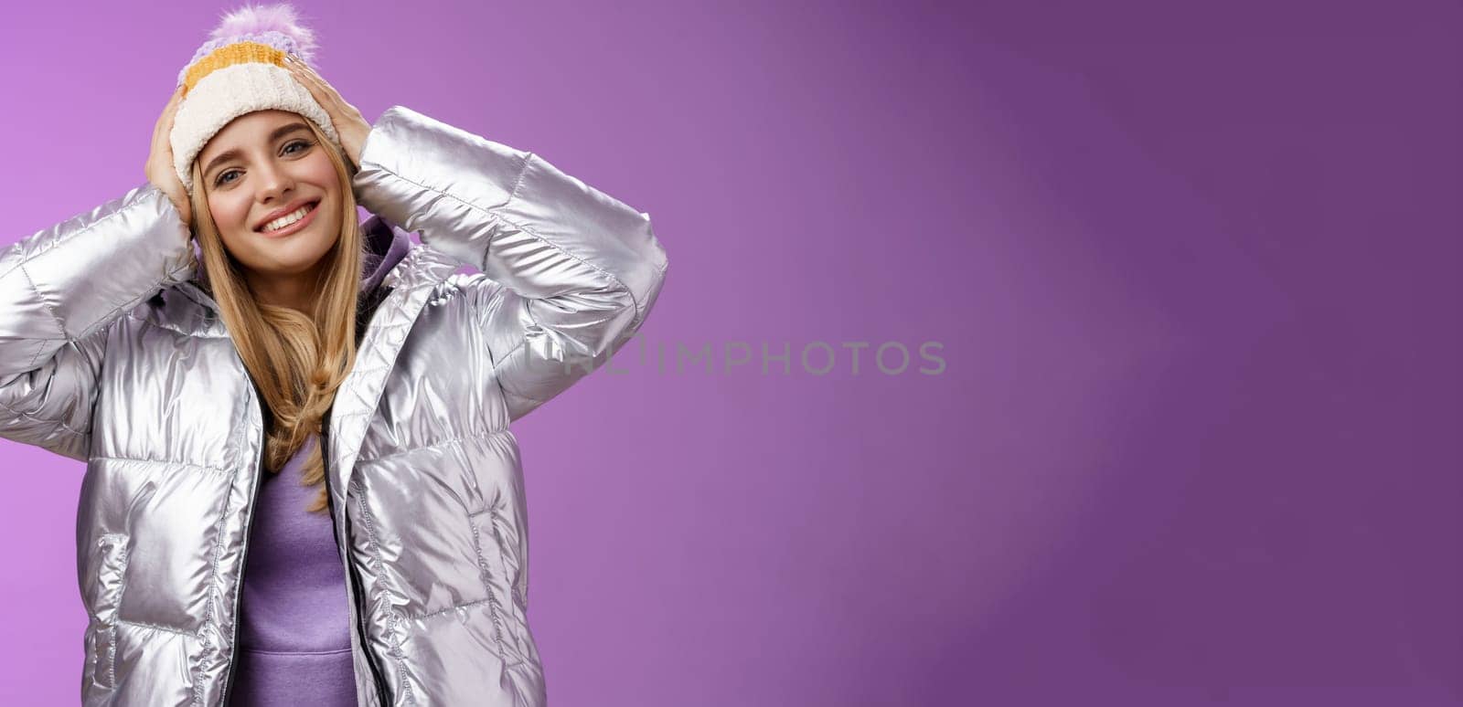 Tender romantic attractive blond female enjoying winter ski resort vacation having fun look pleased smiling broadly tilting head touching hat wearing silver stylish jacket, purple background by Benzoix