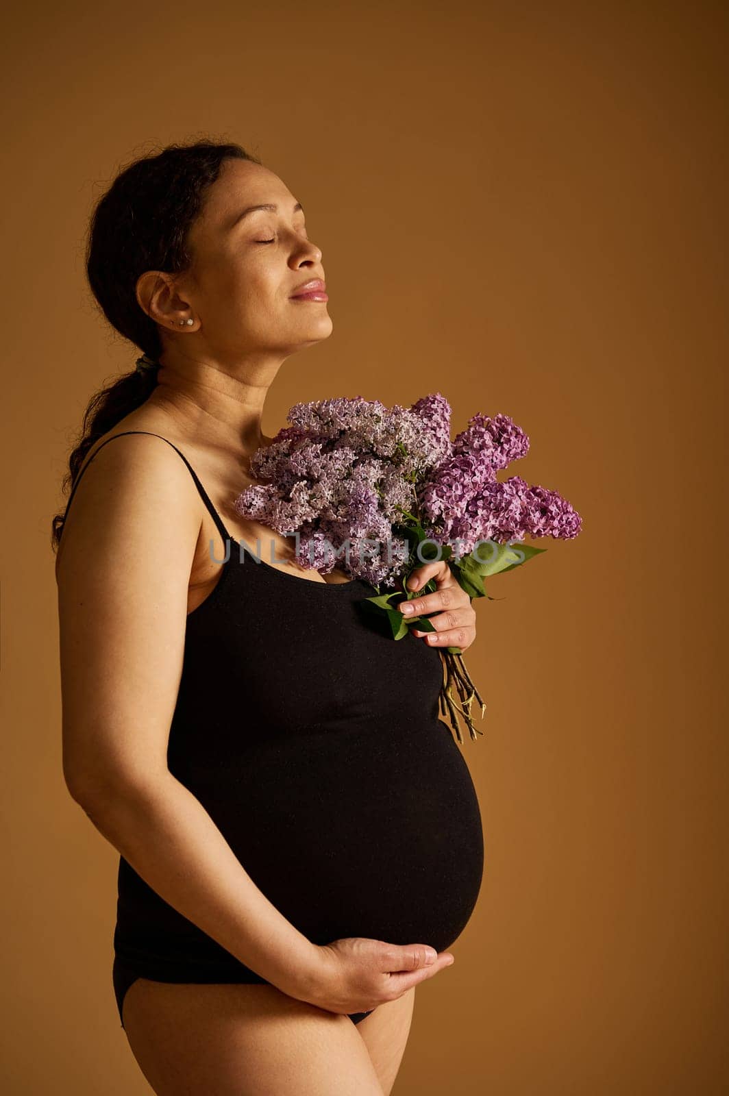 Confident delightful pregnant woman, expectant mother stroking her belly, holding purple lilacs, isolated over beige studio background. Pregnancy. Childbearing. Maternity and parenting concept