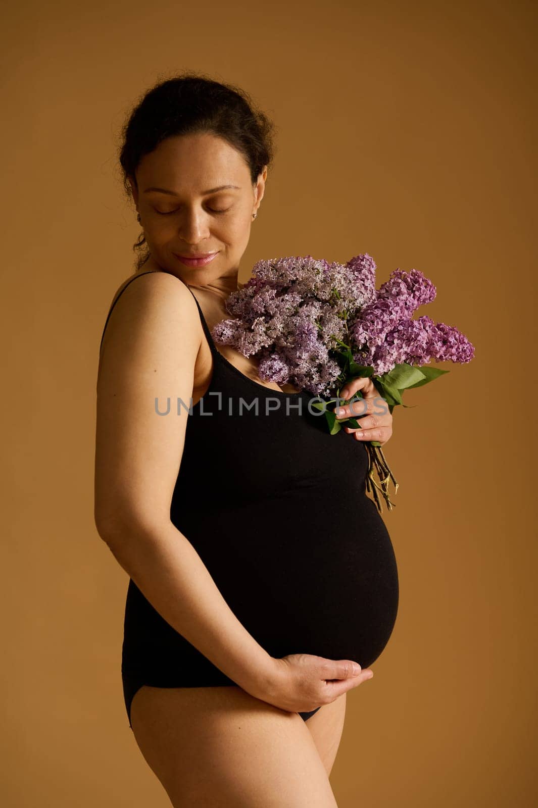 Vertical studio shot of attractive delightful pregnant woman wearing black bodysuit, posing with a bunch of purple lilacs over beige background, enjoying her pregnancy time. Happy maternity concept