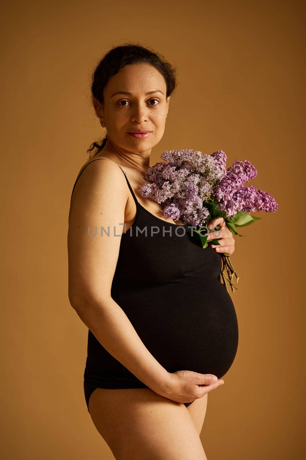 Confident positive pregnant woman in black bodysuit, posing looking at the camera, isolated on beige studio background by artgf