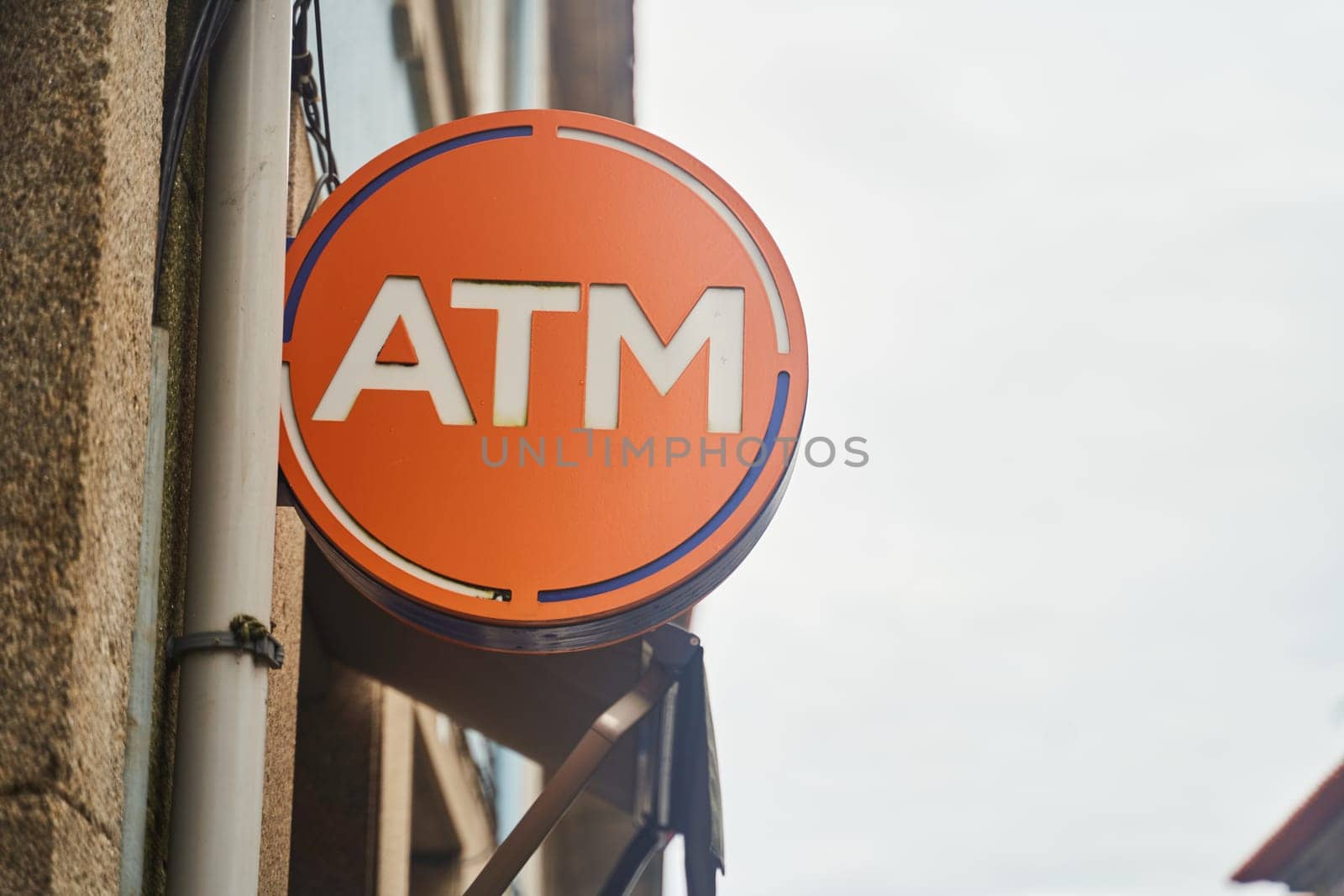 An orange ATM sign with a sky blue logo and metal cylinder on the side of a building. The signage uses a bold font and is in the shape of a circle