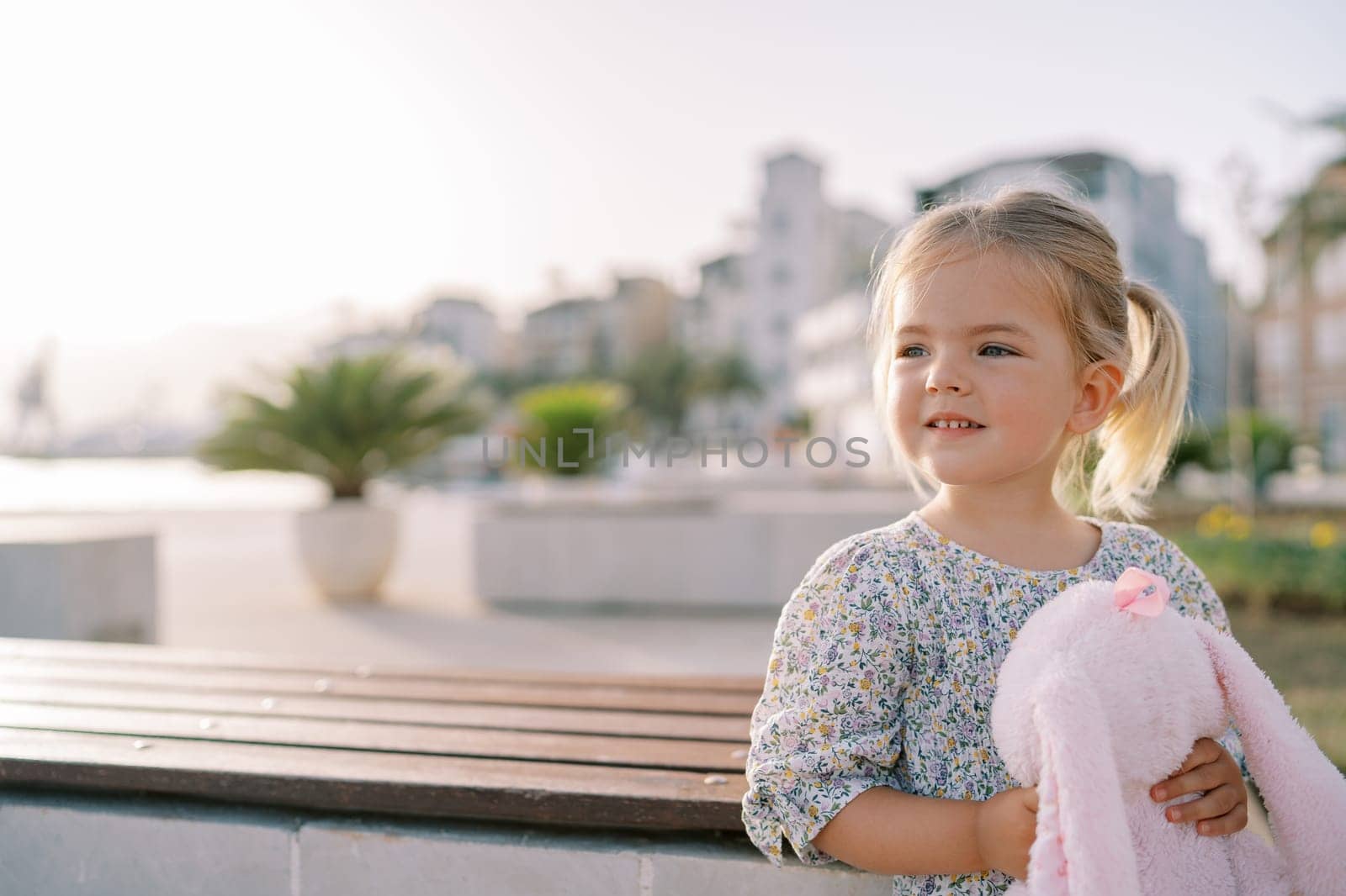 Little smiling girl with a pink soft toy hare stands near a wooden table on the embankment and looks away. High quality photo