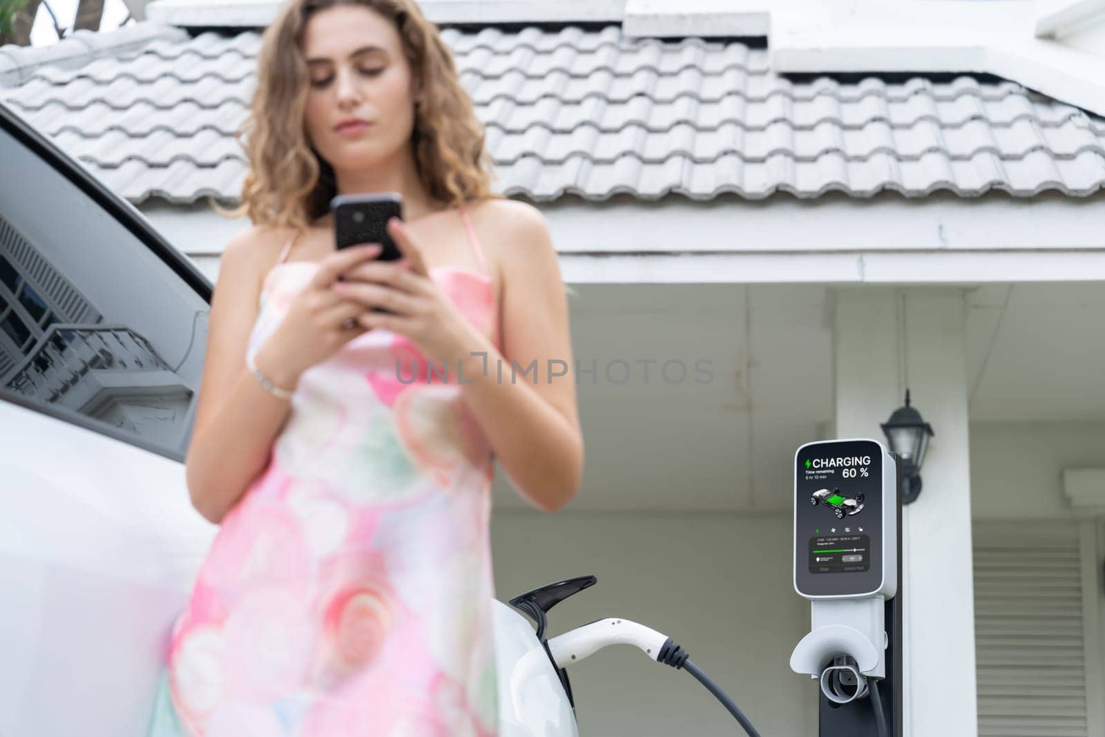 Focused home EV charging station on blurred background of modern eco-friendly woman using smartphone. EV technology utilization for residential home concept. Synchronos