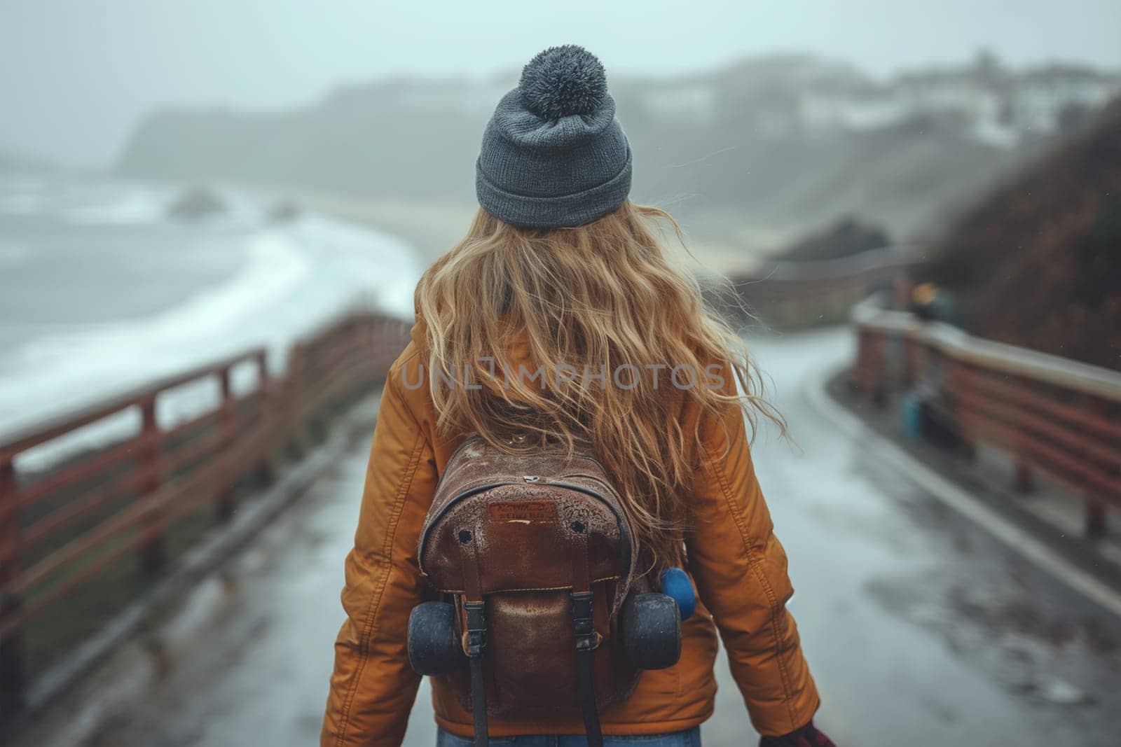 A person bundled up in a warm orange jacket and beanie walks along a foggy seaside pathway, with turbulent ocean waves in the distance.