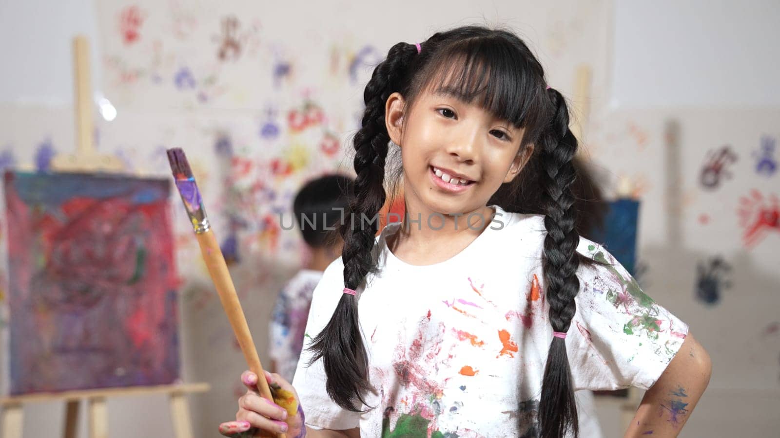 Creative cute girl pose at camera while diverse children painting behind. Young beautiful child looking at camera while standing at stained room and holding paintbrush. Creative activity. Erudition.