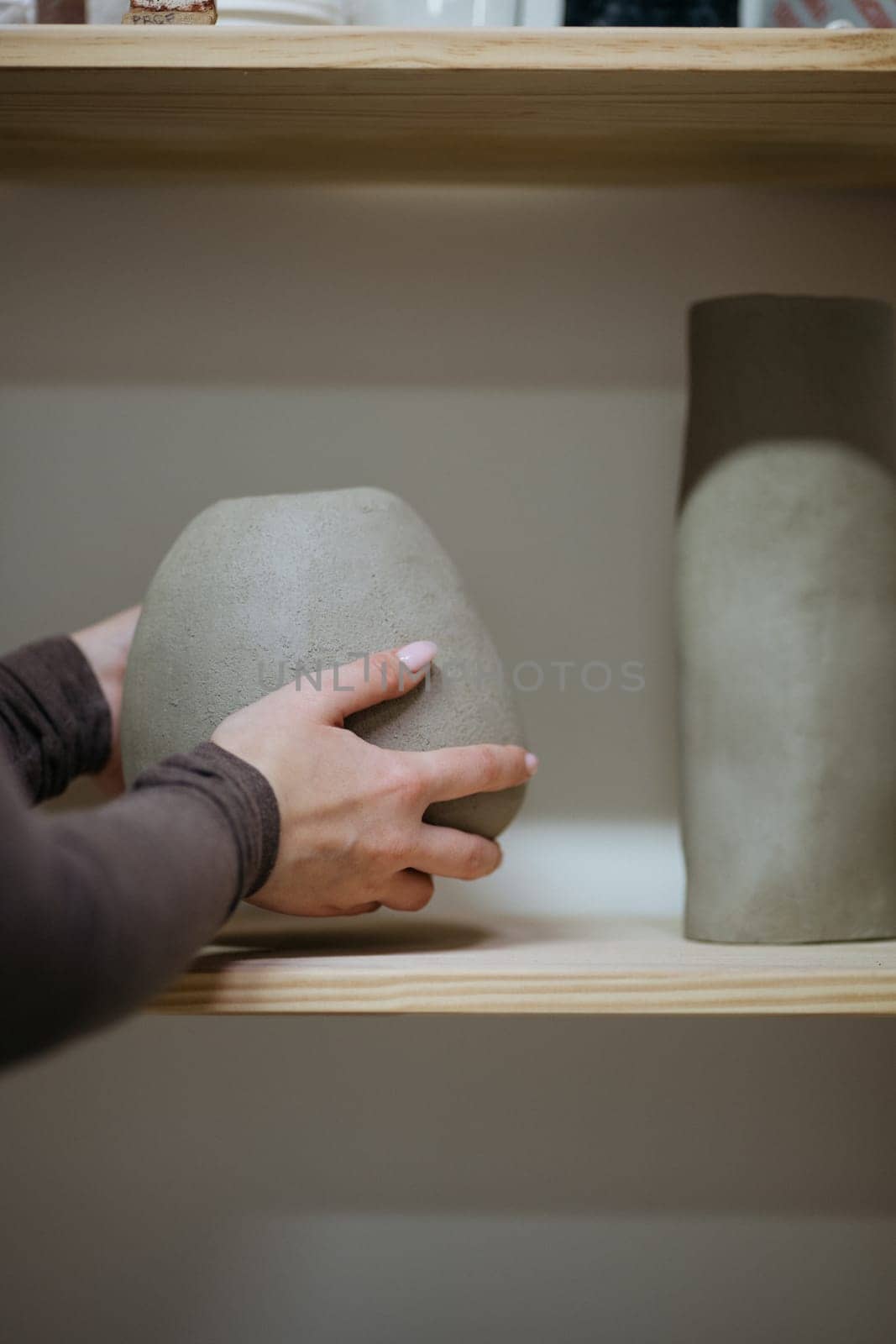 A hand gently grips a sandy-textured ceramic vase on a wooden shelf.