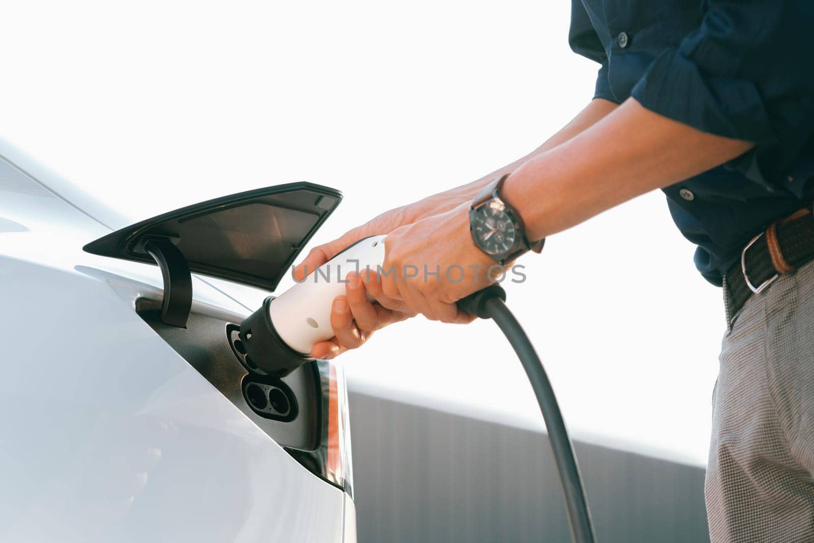 Young man put EV charger to recharge electric car's battery from charging station in city commercial parking lot. Rechargeable EV car for sustainable environmental friendly urban travel. Expedient