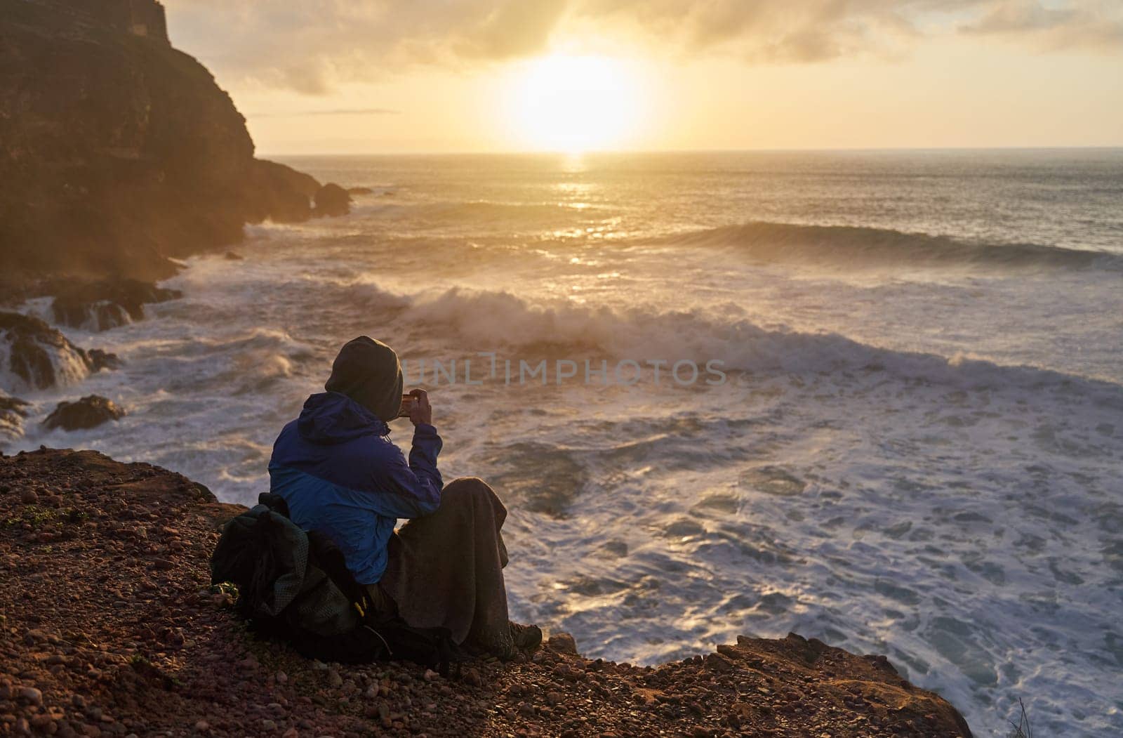 Person sits on cliff, gazing at ocean as sun sets over water, sky, and horizon by driver-s