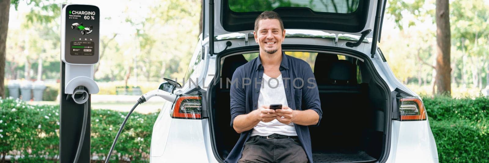 Man using smartphone online banking application to pay for electric car battery charging from EV charging station during vacation road trip at national park or summer forest. Panorama Exalt