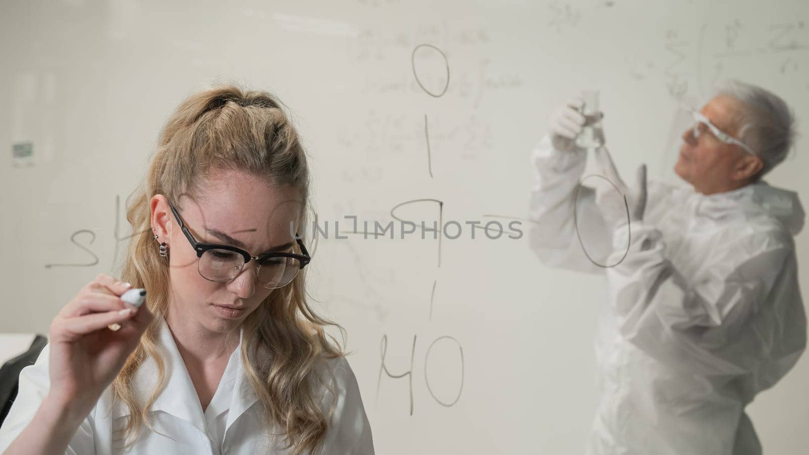 A woman chemist writes a formula on glass. An elderly Caucasian man in a protective suit is doing tests. by mrwed54