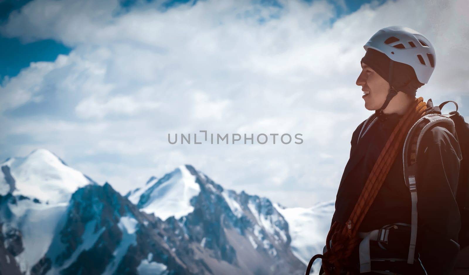 A young man traveler is engaged in mountaineering. In a helmet, with a rope, a harness, gloves, climbs to the top, against the backdrop of a stunning view with snow-capped mountains.