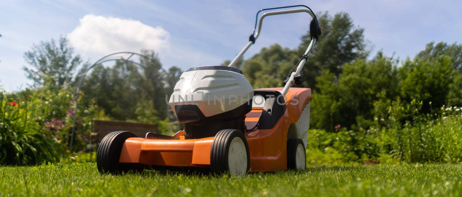 A lawn mower is outside at the beautiful green floral backyard lawn. A lawnmower is cutting a lawn on a summer sunny day.