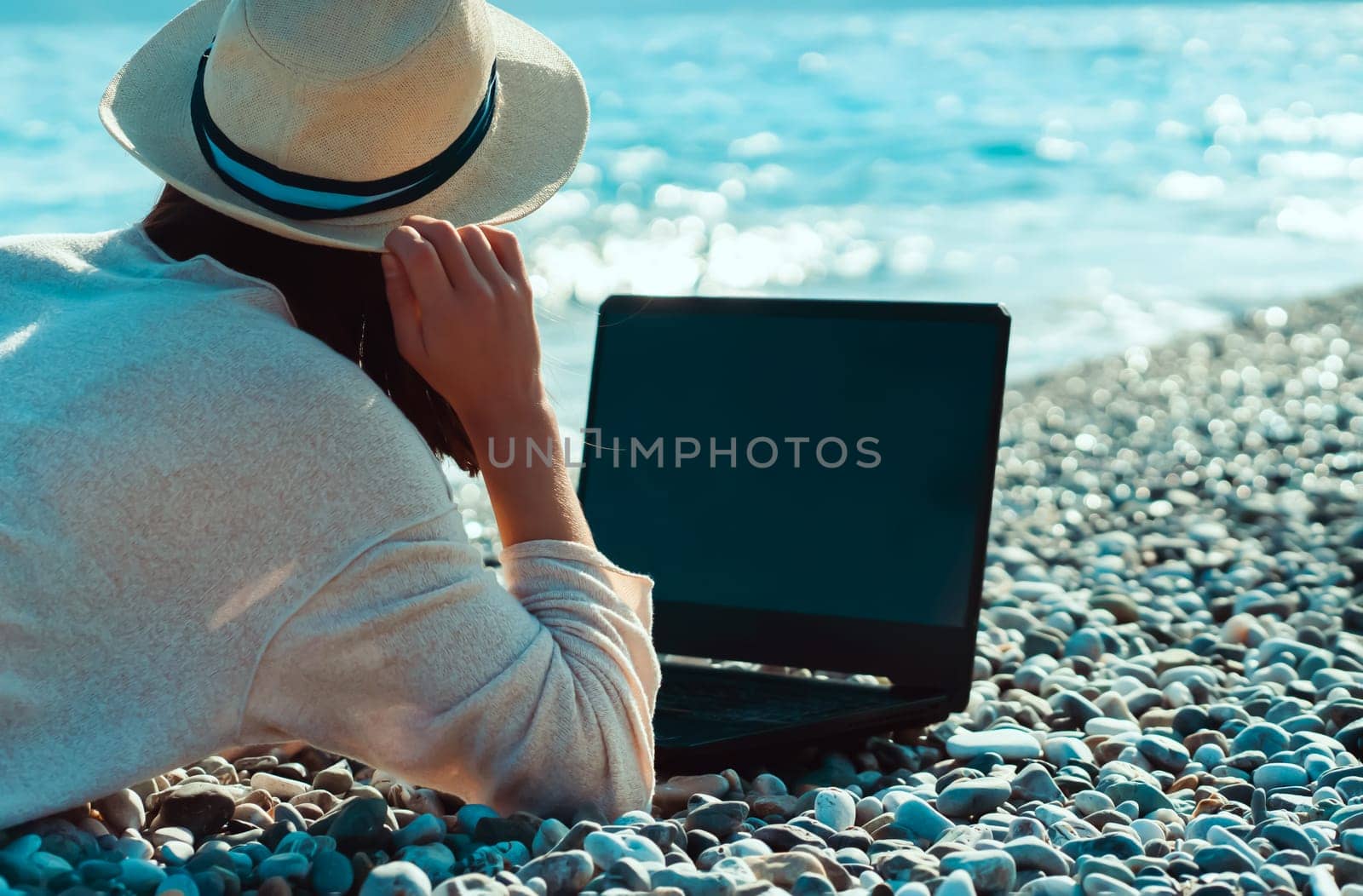 A young girl in a light hat and casual sweater lies on the beach by the sea with a laptop on a sunny day, works, studies, buys tickets during trip, a woman rests on vacation and types on the keyboard.