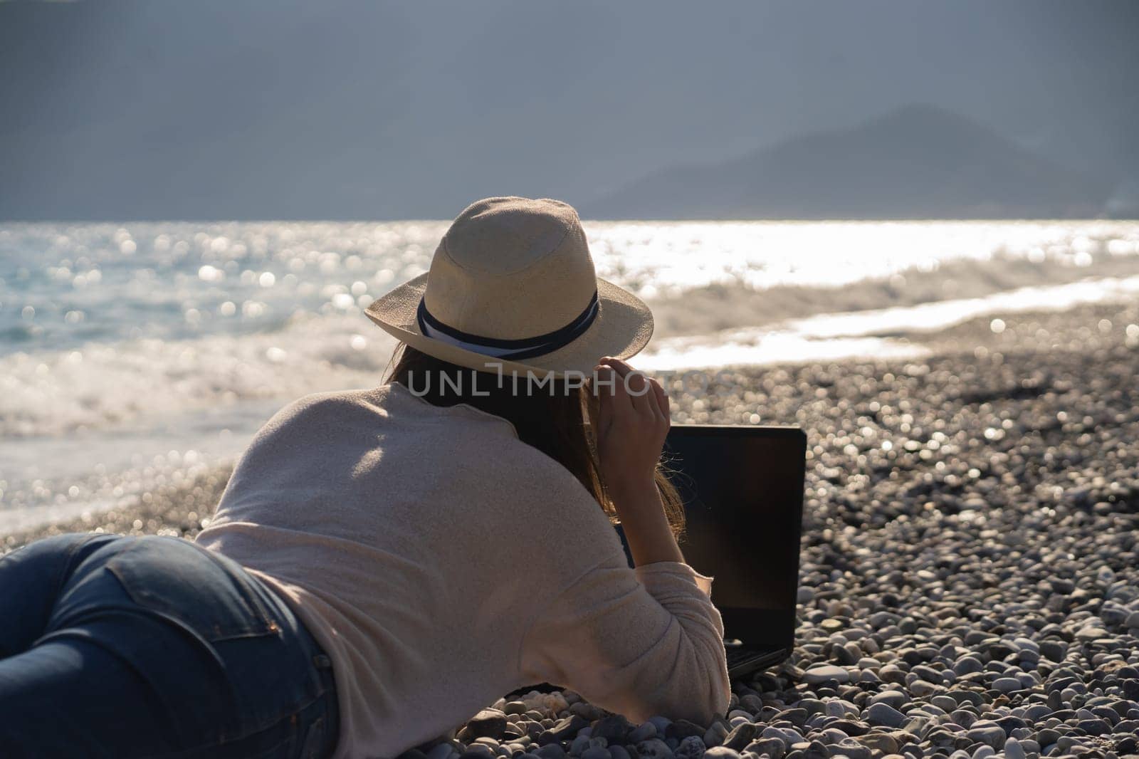 A young girl in a light hat and casual sweater lies on the beach by the sea with a laptop on a sunny day, works, studies, buys tickets during trip, a woman rests on vacation and types on the keyboard.