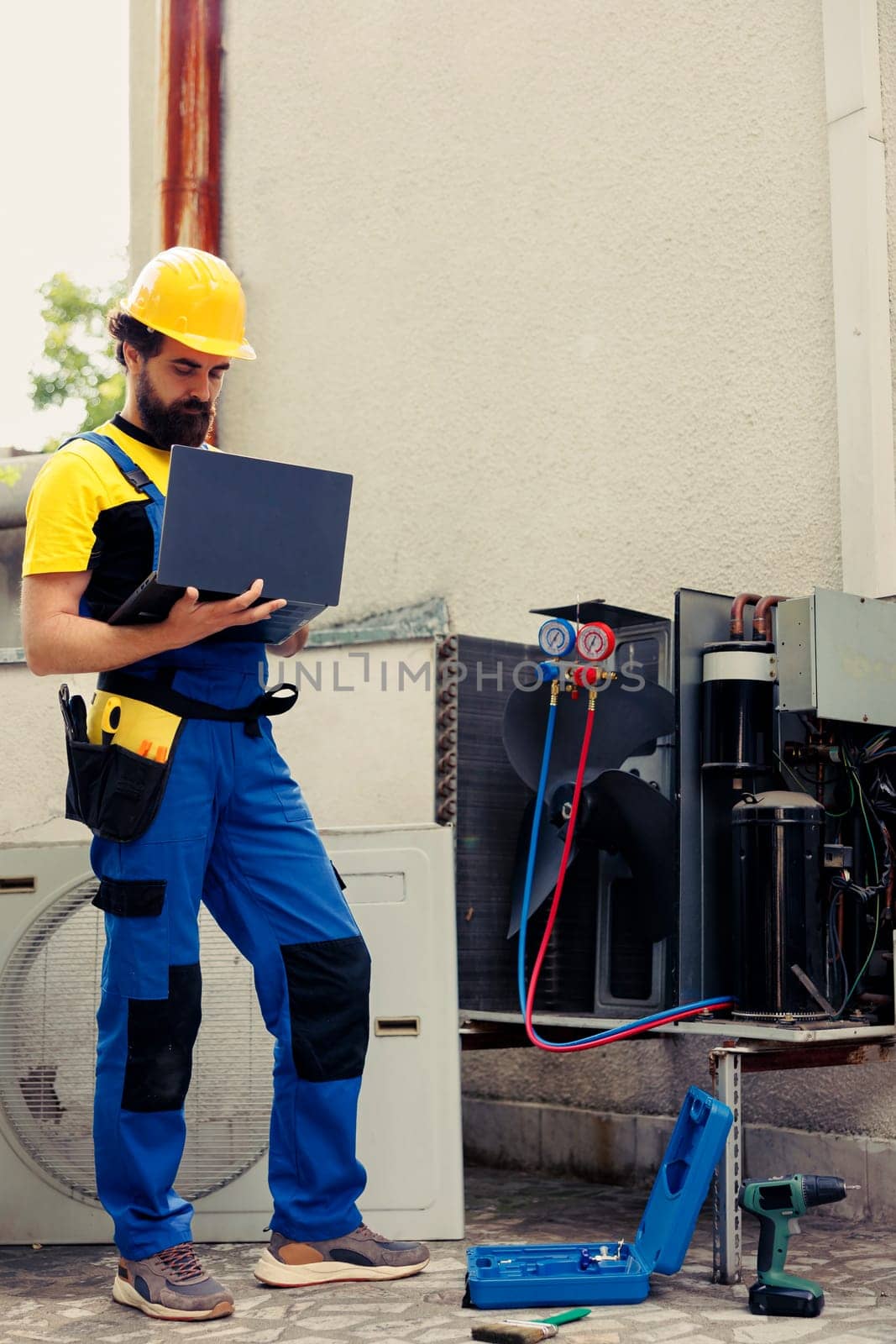 Certified expert commissioned for outside air conditioner routine check up, imputing data on laptop. Trained mechanic doing hvac system safety check, looking for defective internal parts