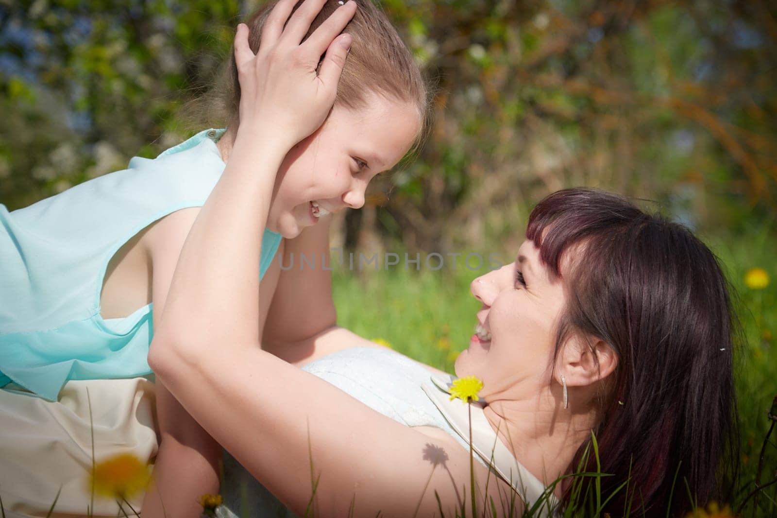 Happy mother and daughter enjoying rest, playing and fun on nature on a green lawn with dandelions and blooming apple tree on the background. Woman and girl resting outdoors in summer and spring day by keleny