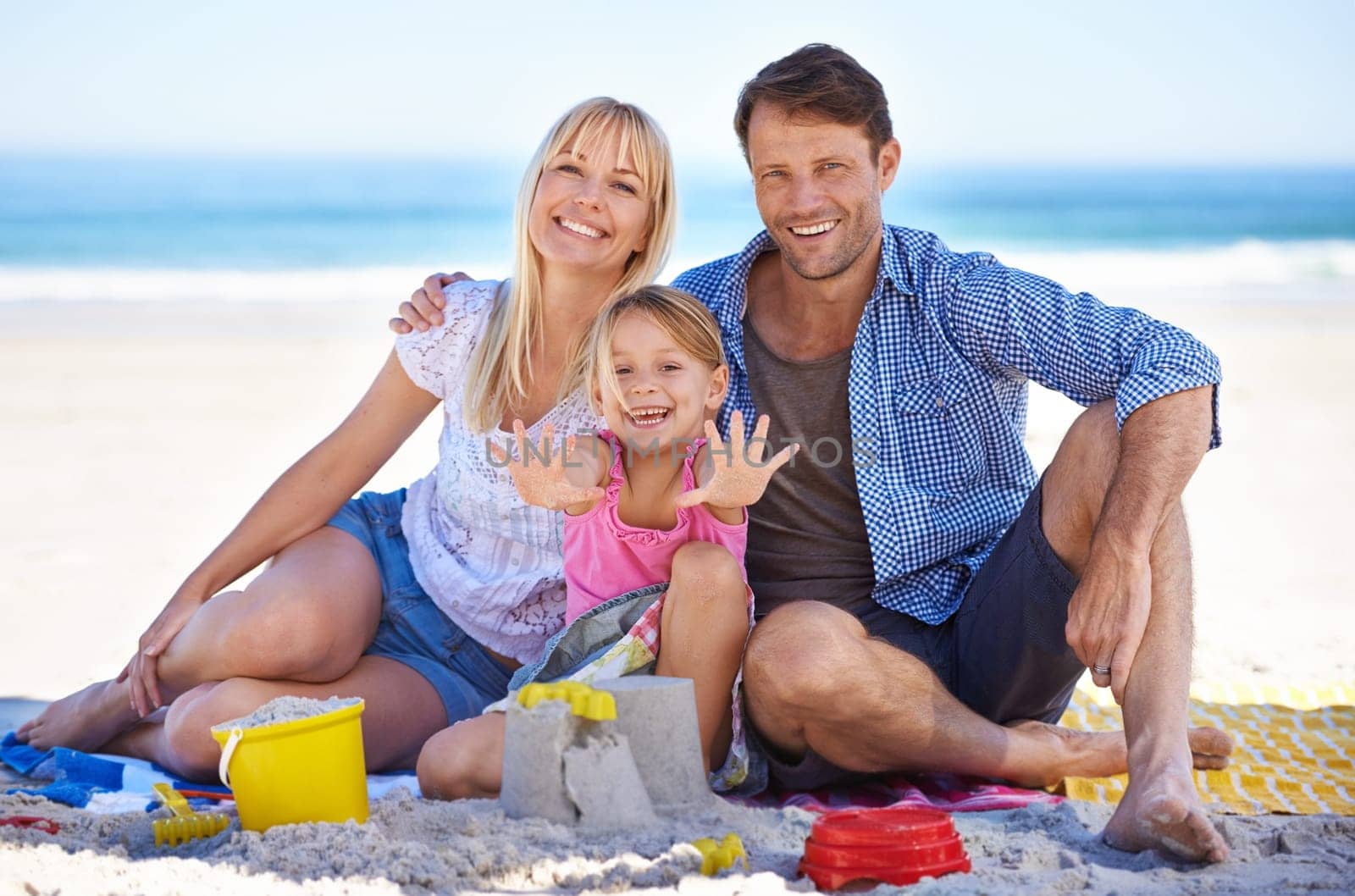 Parents, girl and sandcastle in portrait at beach, blanket and excited with hug for holiday in summer. Father, mother and daughter with picnic by ocean for vacation in nature with love in sunshine.