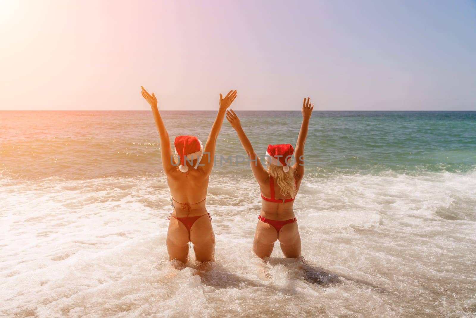 Women Santa hats ocean play. Seaside, beach daytime, enjoying beach fun. Two women in red swimsuits and Santa hats are enjoying themselves in the ocean, kneeling in the waves and raising their hands up