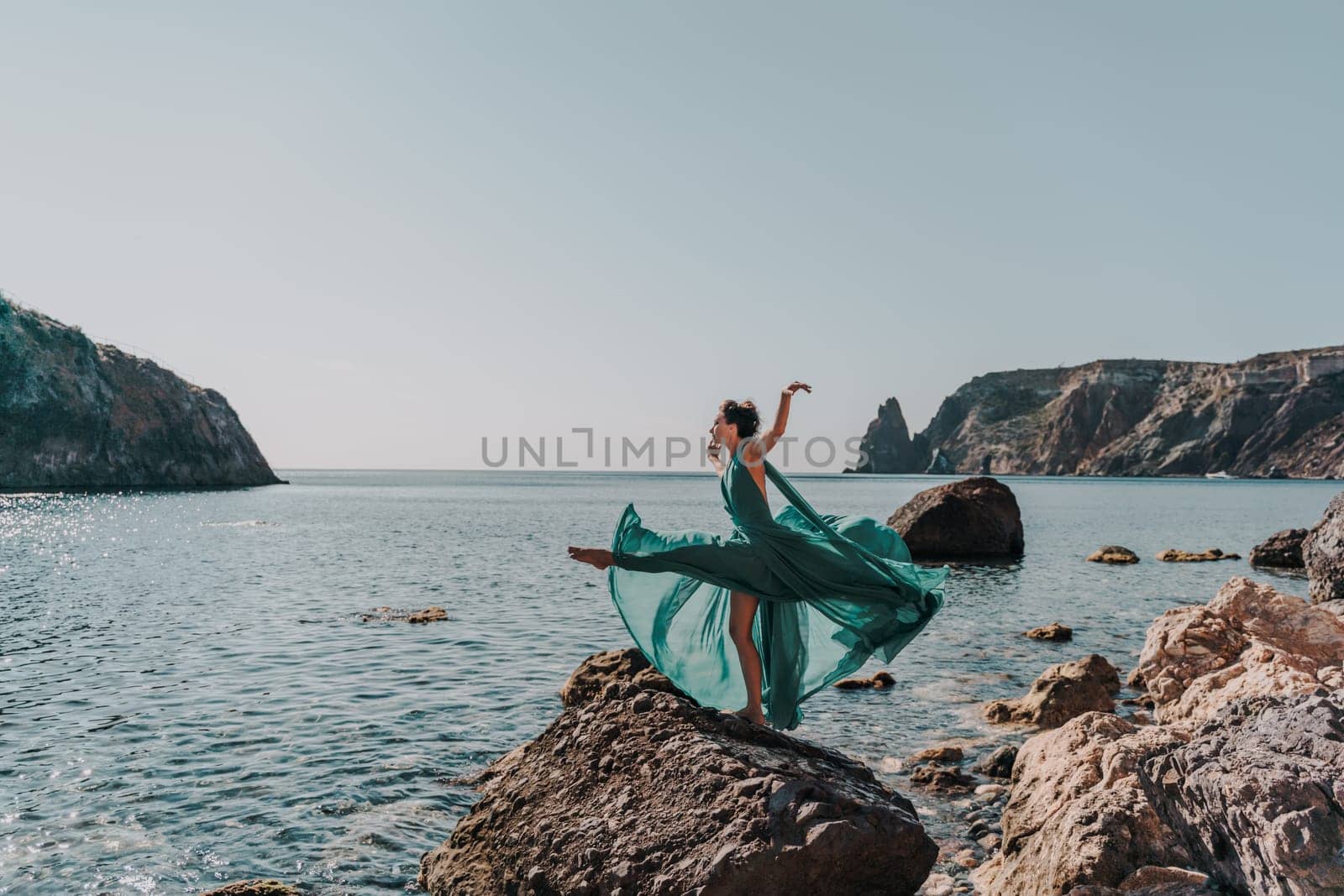 Woman green dress sea. Female dancer in a long mint dress posing on a beach with rocks on sunny day. Girl on the nature on blue sky background