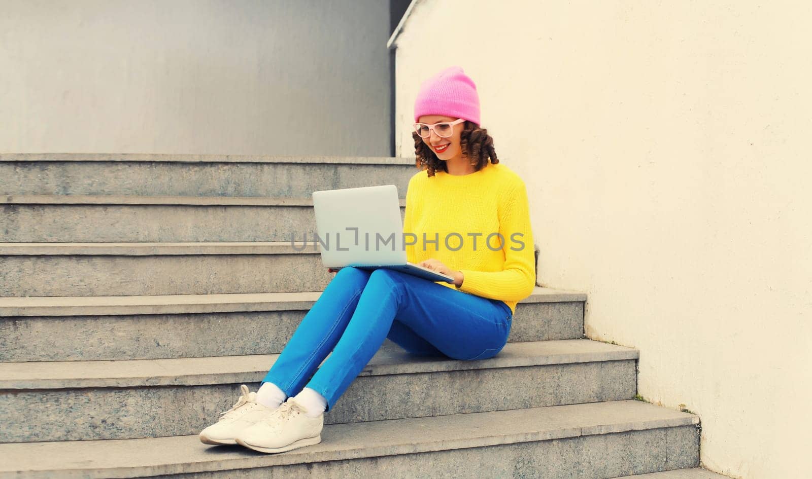 Portrait of stylish modern young woman working with laptop wearing a colorful clothes, eyeglasses in the city