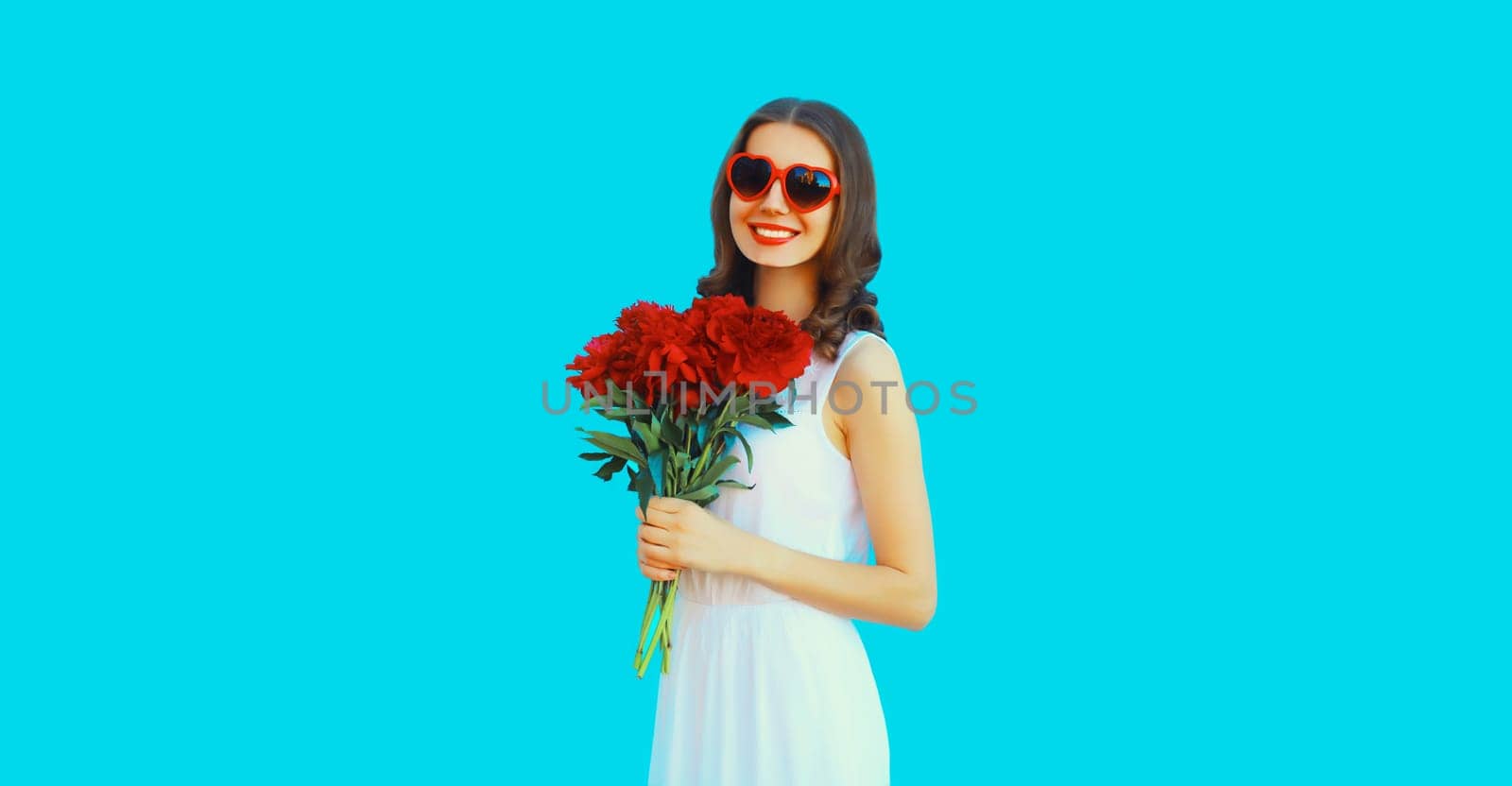 Portrait of beautiful smiling woman with bouquet of red rose flowers in heart shaped sunglasses on blue background