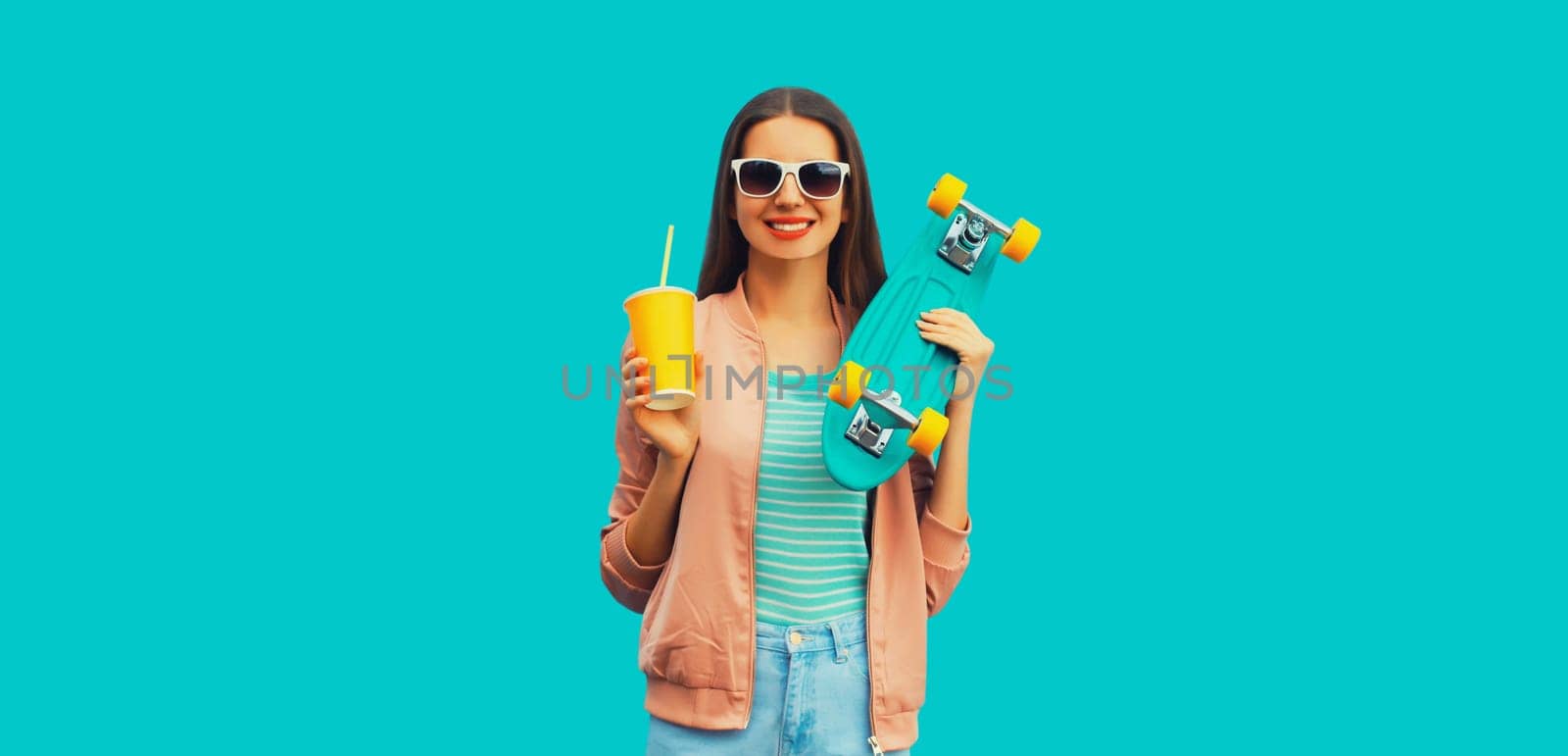 Portrait of stylish young woman with skateboard drinking juice on blue background