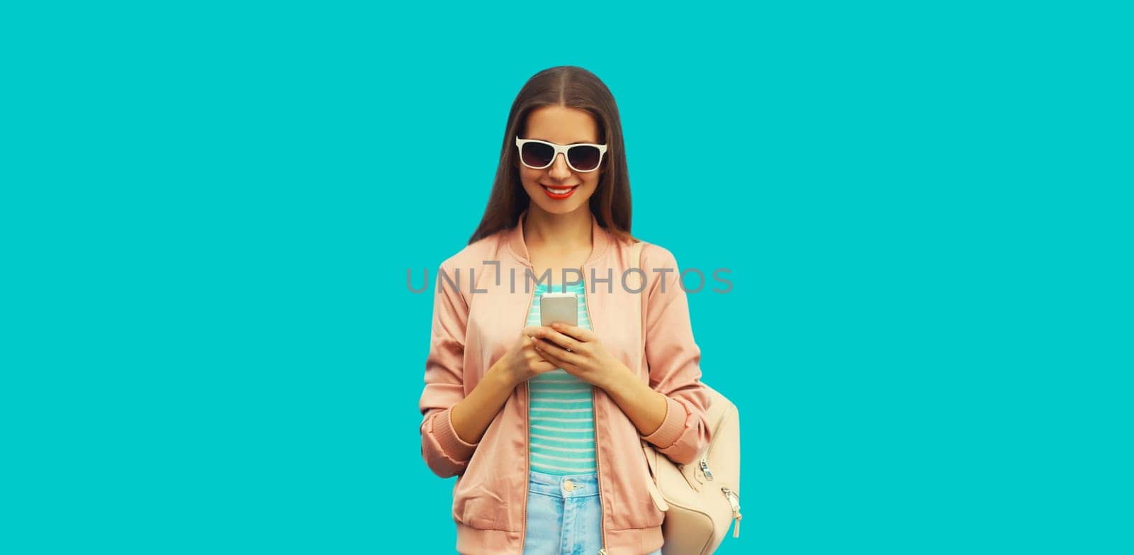 Portrait of smiling young woman with smartphone wearing backpack and sunglasses on blue background