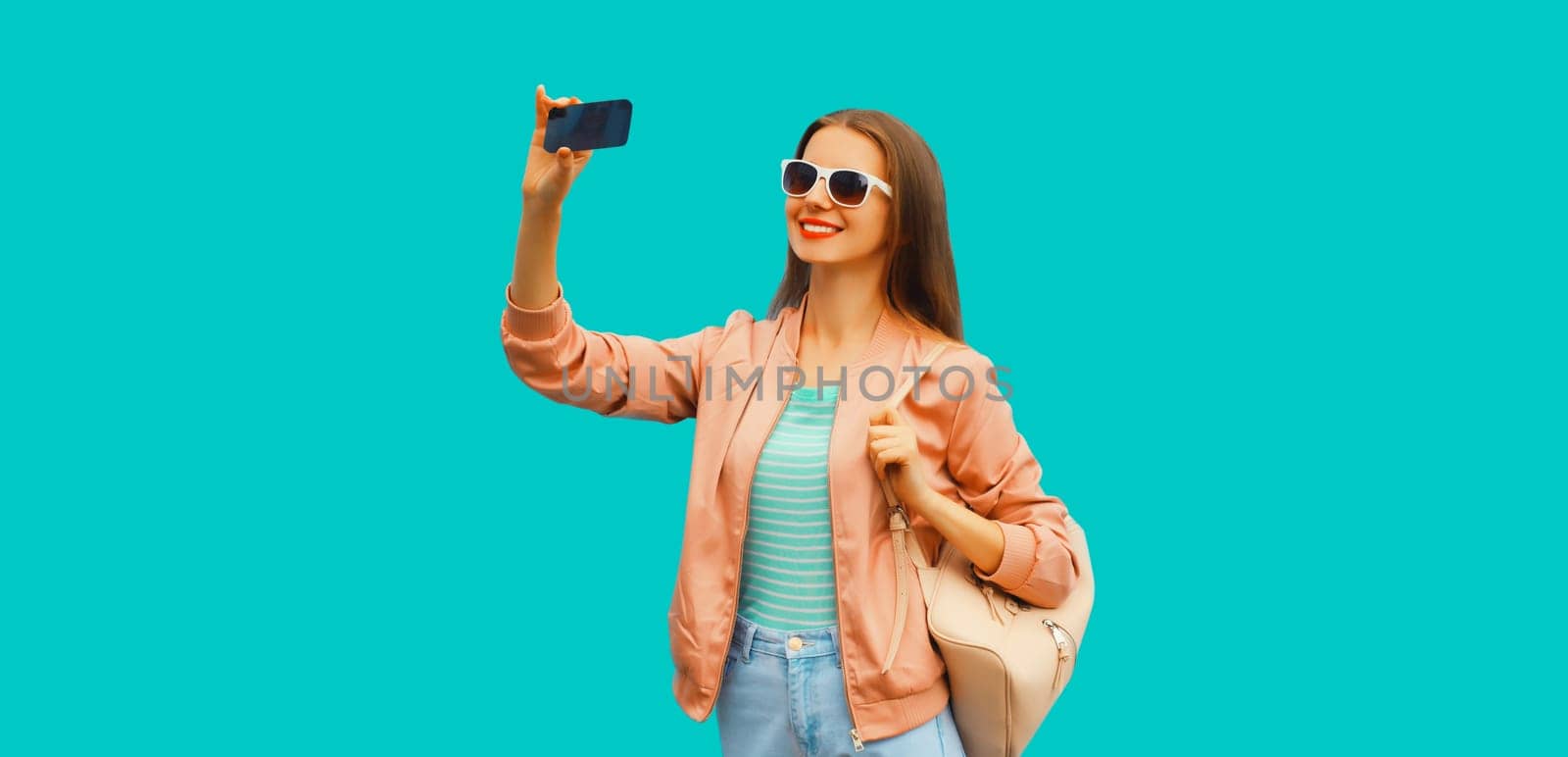 Portrait of happy smiling young woman taking selfie with smartphone wearing backpack on blue background