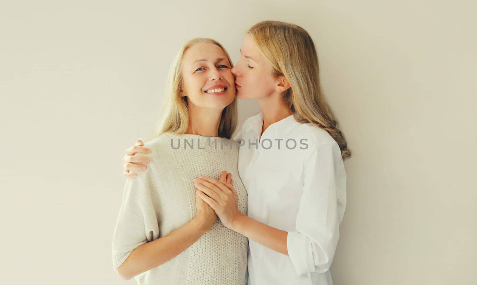 Portrait of adult daughter kissing her happy smiling caucasian middle aged mother on studio background