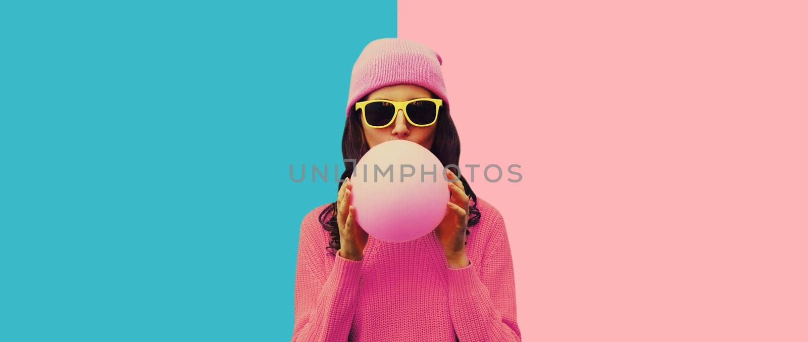 Fashion portrait of stylish cool young woman inflating chewing gum or balloon in pink hat, sunglasses on colorful studio background