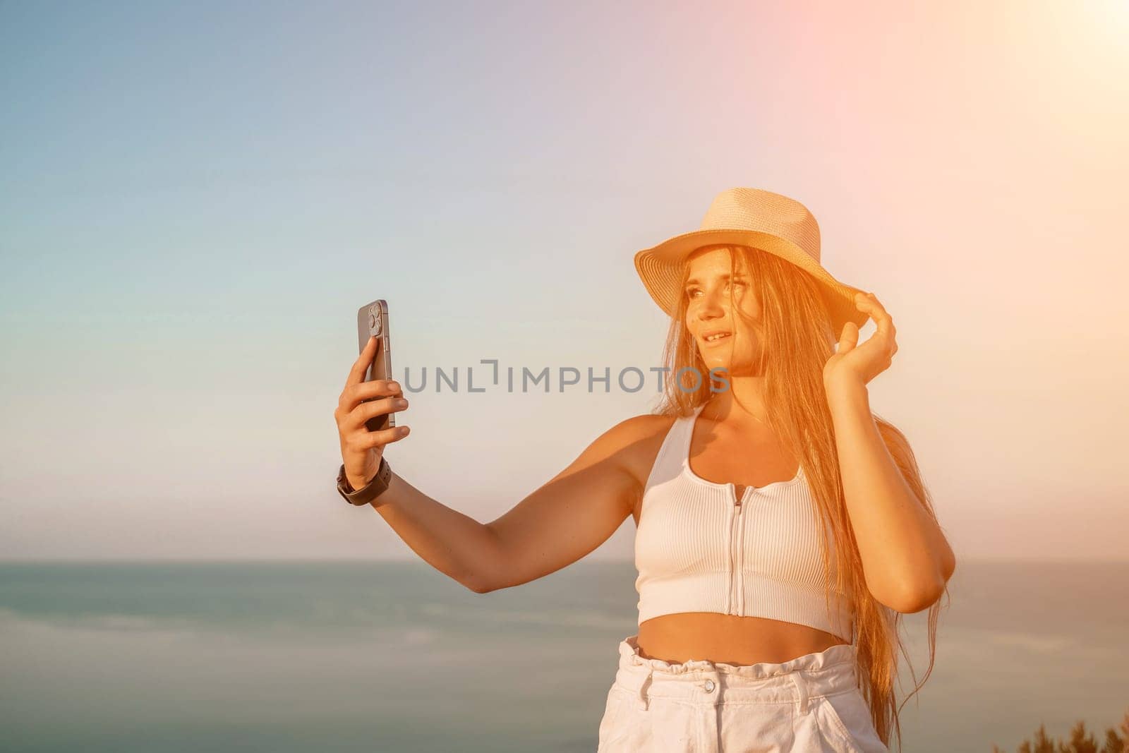Selfie woman in a hat, white tank top, and shorts captures a selfie shot with her mobile phone against the backdrop of a serene beach and blue sea
