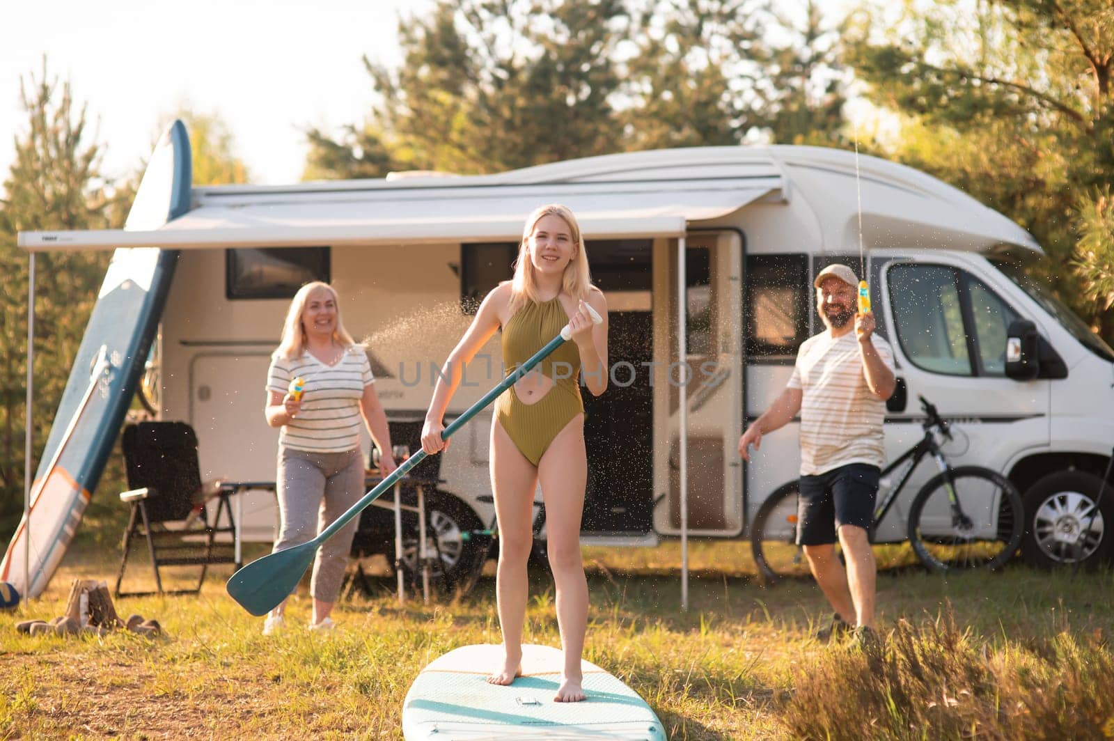 the family is resting next to their mobile home. My daughter is standing with a paddle on a sup board, and her parents pour water on her.