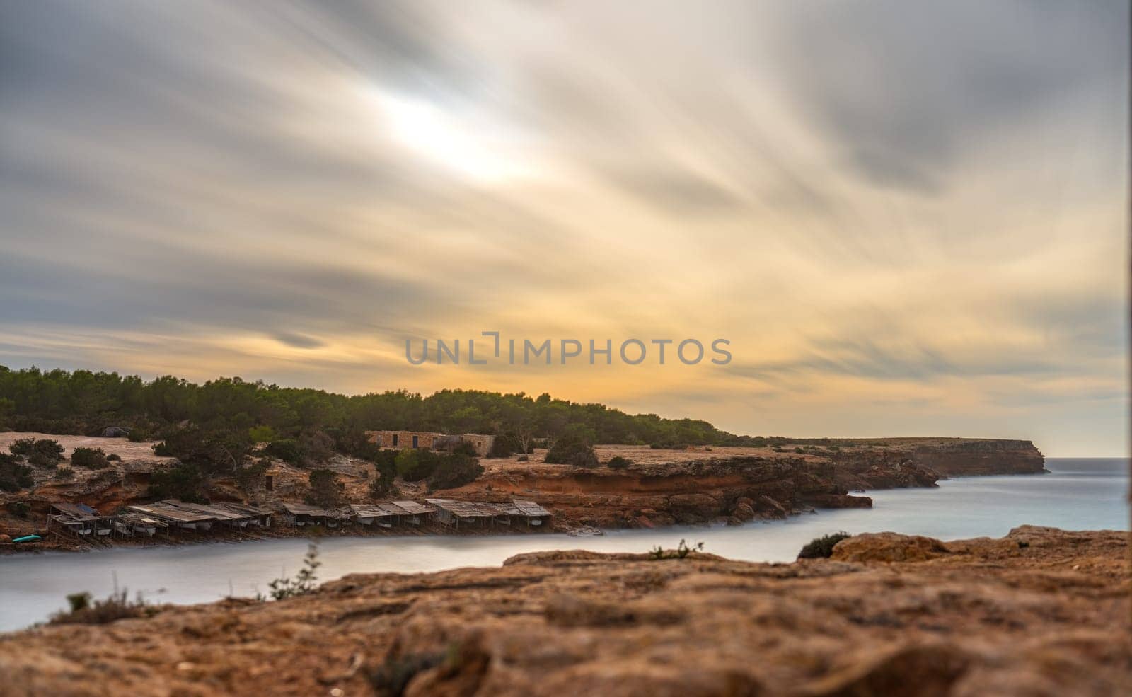 Long exposure photo captures rocky cove in Formentera with fishermen's boats, smooth sea, and fluffy clouds at sunset.