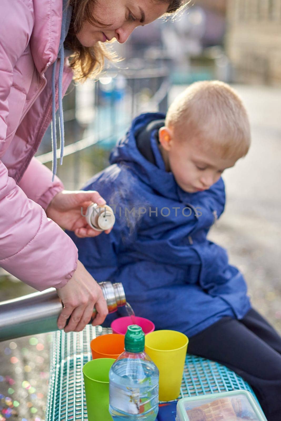 Family Picnic Delight: Cheerful 8-Year-Old Blond Boy in Blue Winter Jacket Sits on Bench While Mom Pours Tea from Thermos, Autumn or Winter. Immerse yourself in the warmth of family moments with this heartening image featuring a joyful 8-year-old blond boy in a blue winter jacket, sitting on a bench while his mom pours tea from a thermos into colorful plastic cups. The photograph captures the essence of a cozy family picnic in the refreshing outdoors during the autumn or winter season.